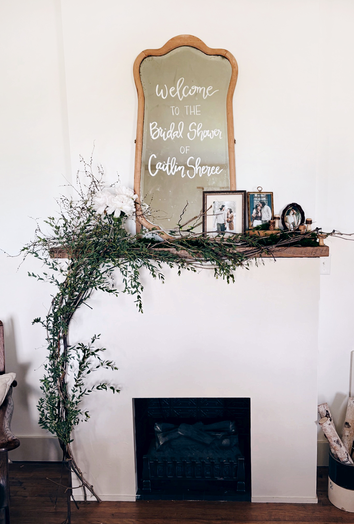 fireplace with old mirror on mantel that says welcome to bridal shower with greenery and old frames on mantel